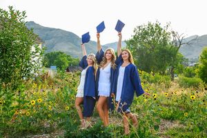 Graduates in blue caps and gowns celebrate by throwing their mortarboards into the air in a flower-filled meadow with mountains.