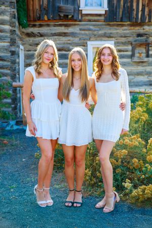Three people in white dresses pose together in front of a rustic cabin exterior.