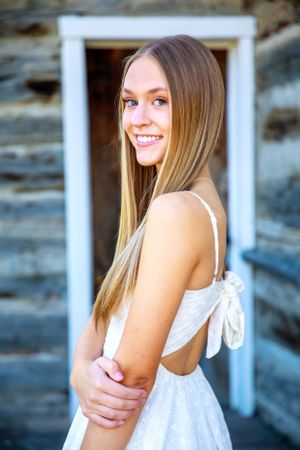 Person in white dress with open back poses by rustic cabin with wooden logs and white doorframe.
