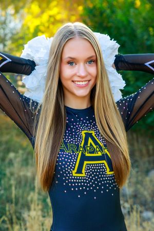 Cheerful person in dark cheer uniform with white pom poms poses outdoors in autumn setting.