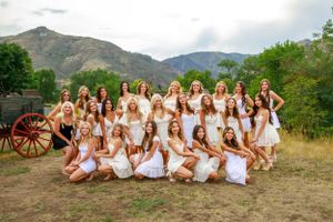Large group of women in white dresses pose together in mountain setting with vintage wagon.