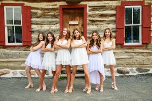 Women in white dresses pose playfully outside rustic cabin with red shutters.