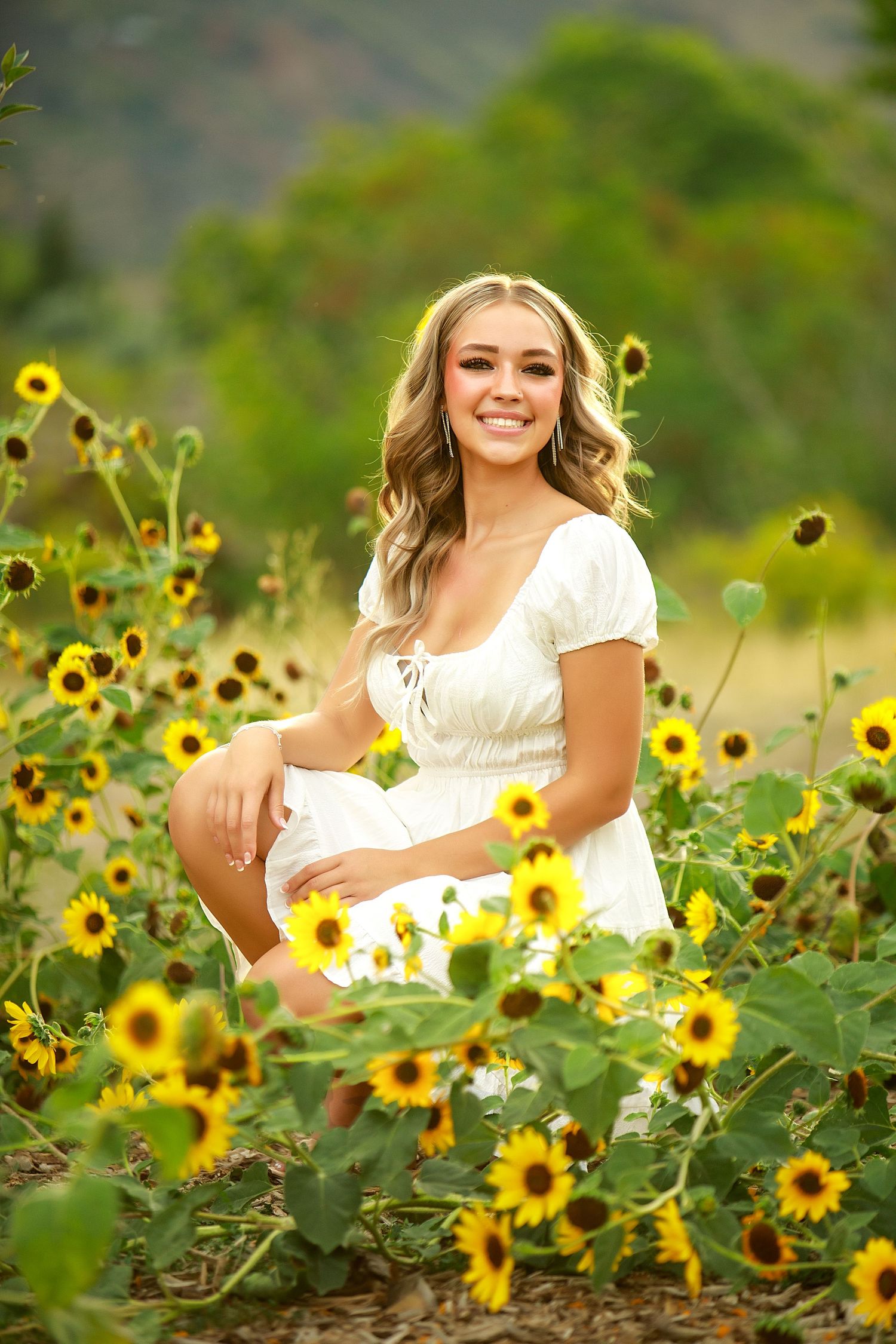 Woman in white dress poses in field of bright yellow sunflowers with mountains in background.