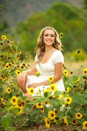 Woman in white dress poses in field of bright yellow sunflowers with mountains in background.