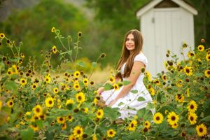 Woman in white dress sits among blooming sunflowers with small white shed in background.