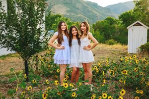 Three women in white summer dresses pose together in a mountain garden filled with bright yellow sunflowers.