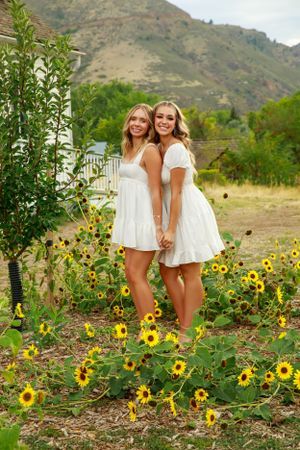 Two women in white sundresses pose together in sunflower field with mountains behind them.