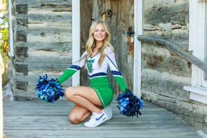Cheerleader poses for senior photos on rustic stone steps wearing blue and white uniform with pom poms.