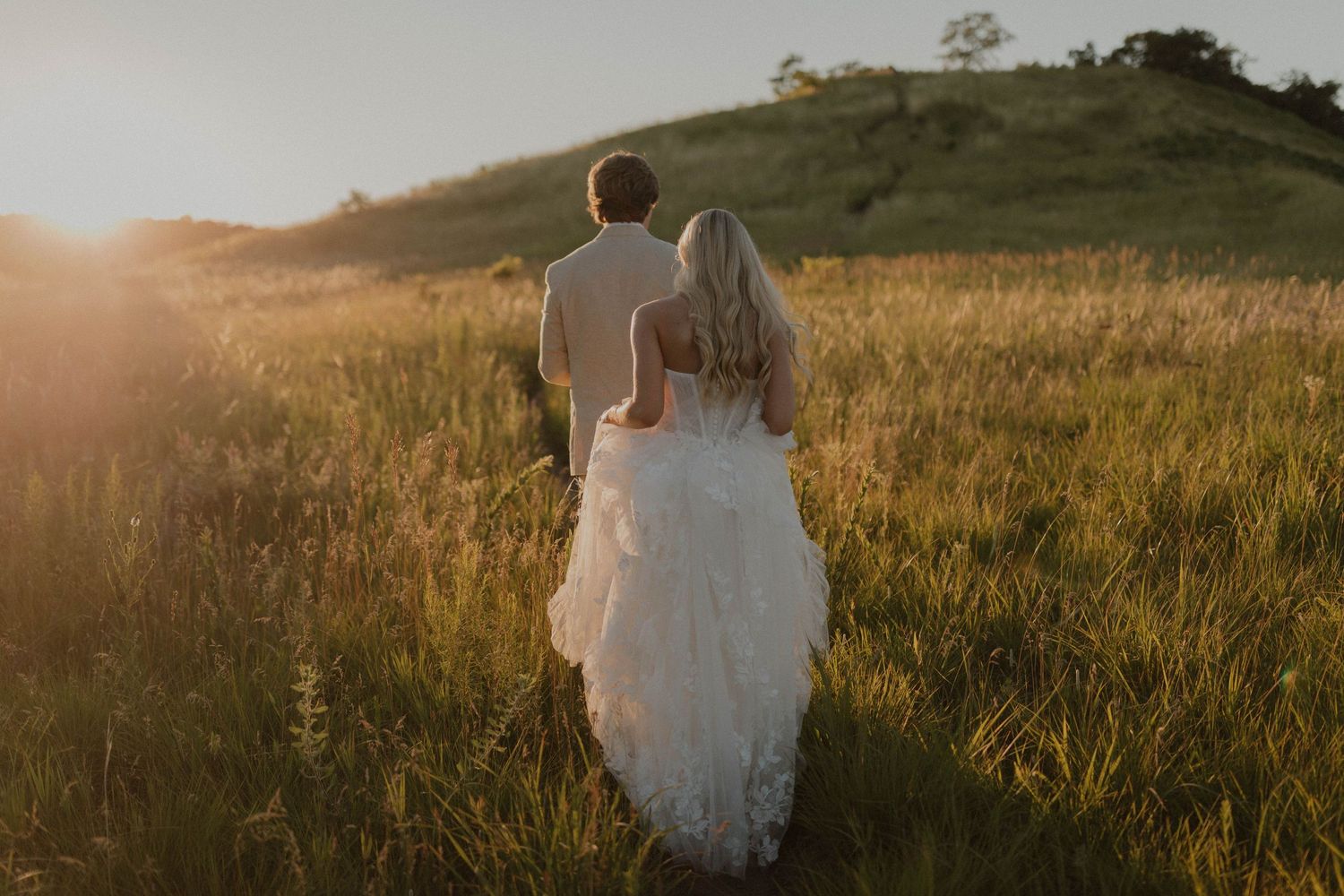 Couple walking through a golden field at sunset holding hands in romantic wedding attire.