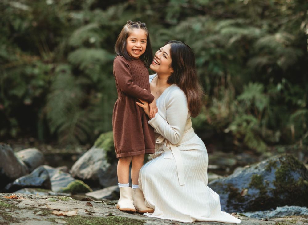 A parent and child share a tender moment while kneeling on rocks by a stream in a lush forest setting.