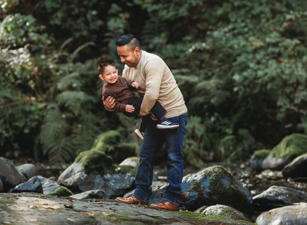A parent and child share a playful moment while balancing on rocks by a stream in a lush forest setting.