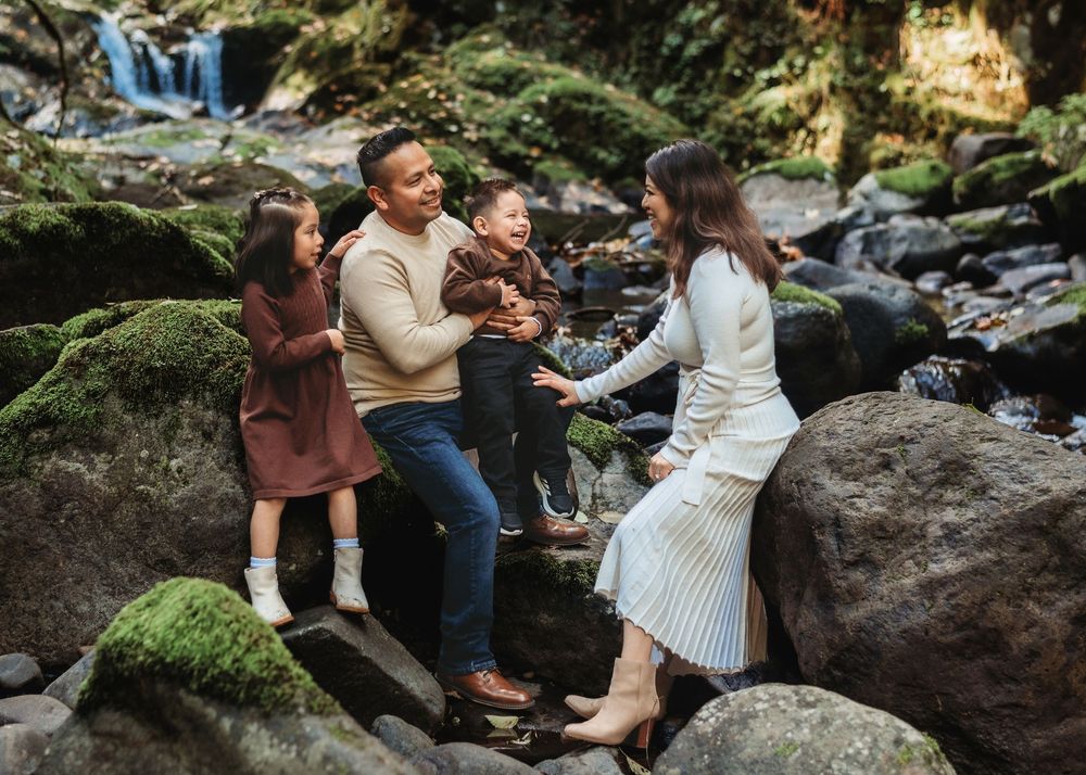 A couple sits together on mossy rocks near a cascading waterfall in a peaceful forest setting.
