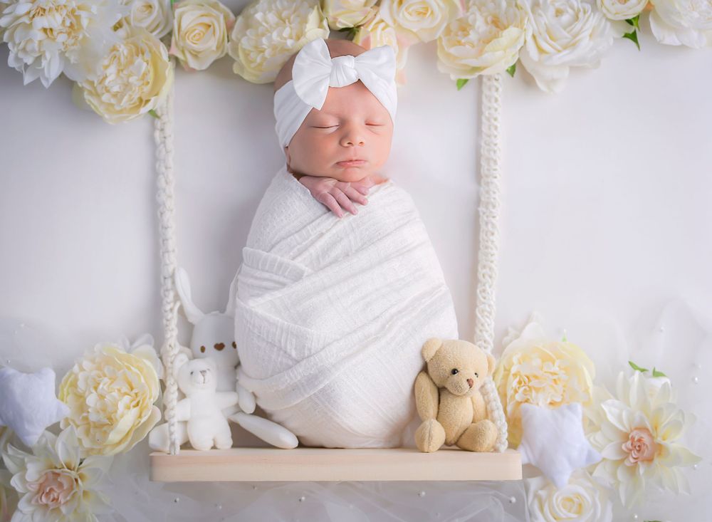 Newborn baby girl on swing prop with cream, white flowers, soft studio lighting by Nicola in Woodside creating timeless gentle portrait.