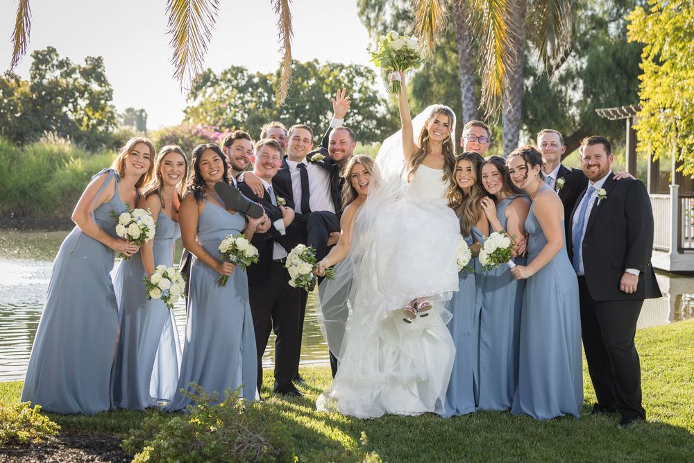 Wedding party posing outdoors near a lake, bridesmaids in blue dresses, groomsmen in suits, bride lifted by groom.