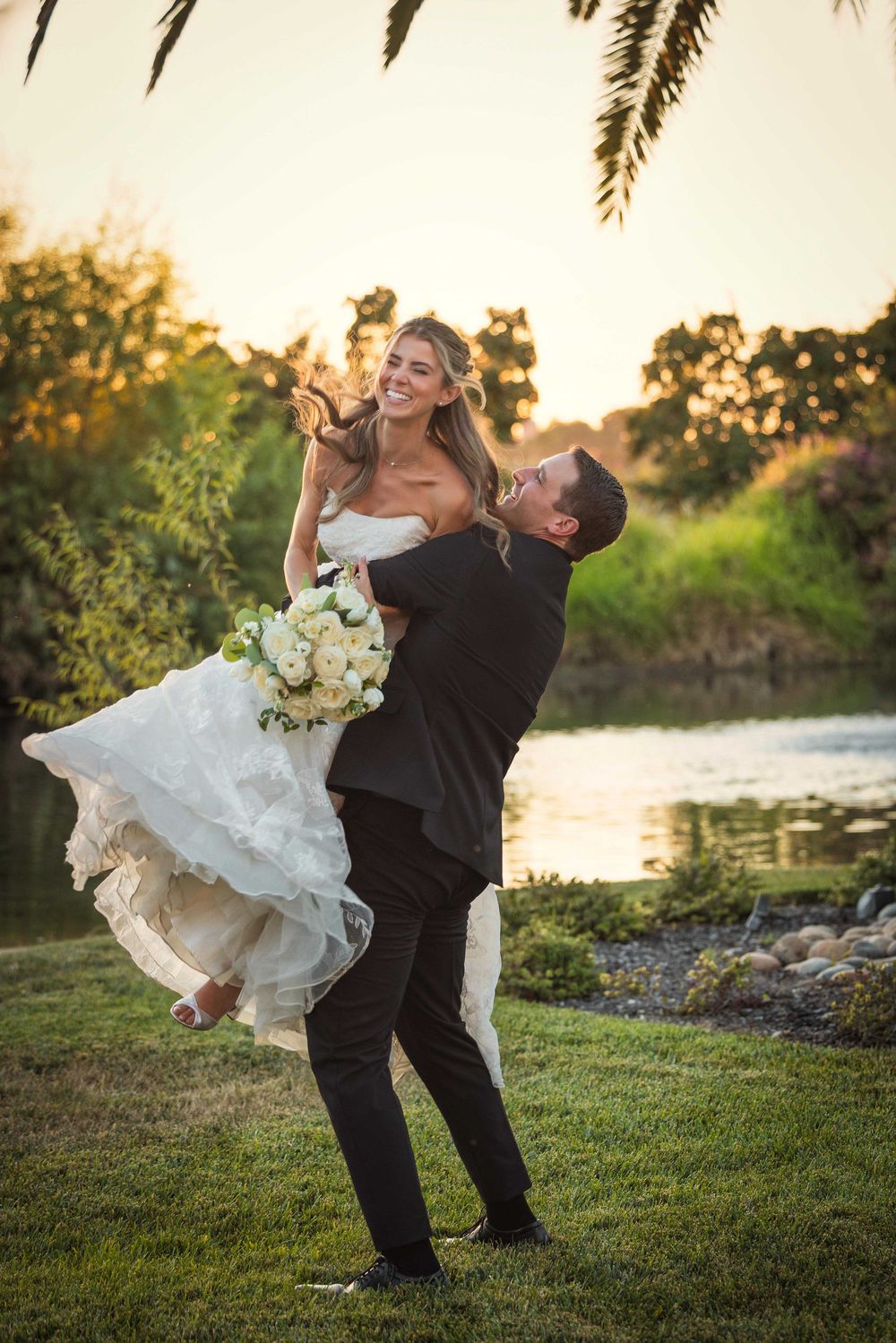 Groom lifts smiling bride in white gown outdoors at sunset near a pond, surrounded by lush greenery.
