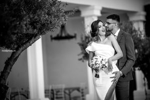 Elegant black and white wedding photo showing a couple embracing outdoors near a Mediterranean style building.