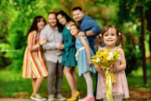 A child in pink dress holds bright yellow flowers in a park while others stand blurred in the background.