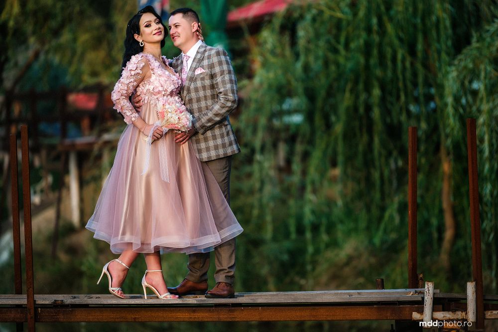 A couple embraces on a wooden deck with lush willow trees in the background.