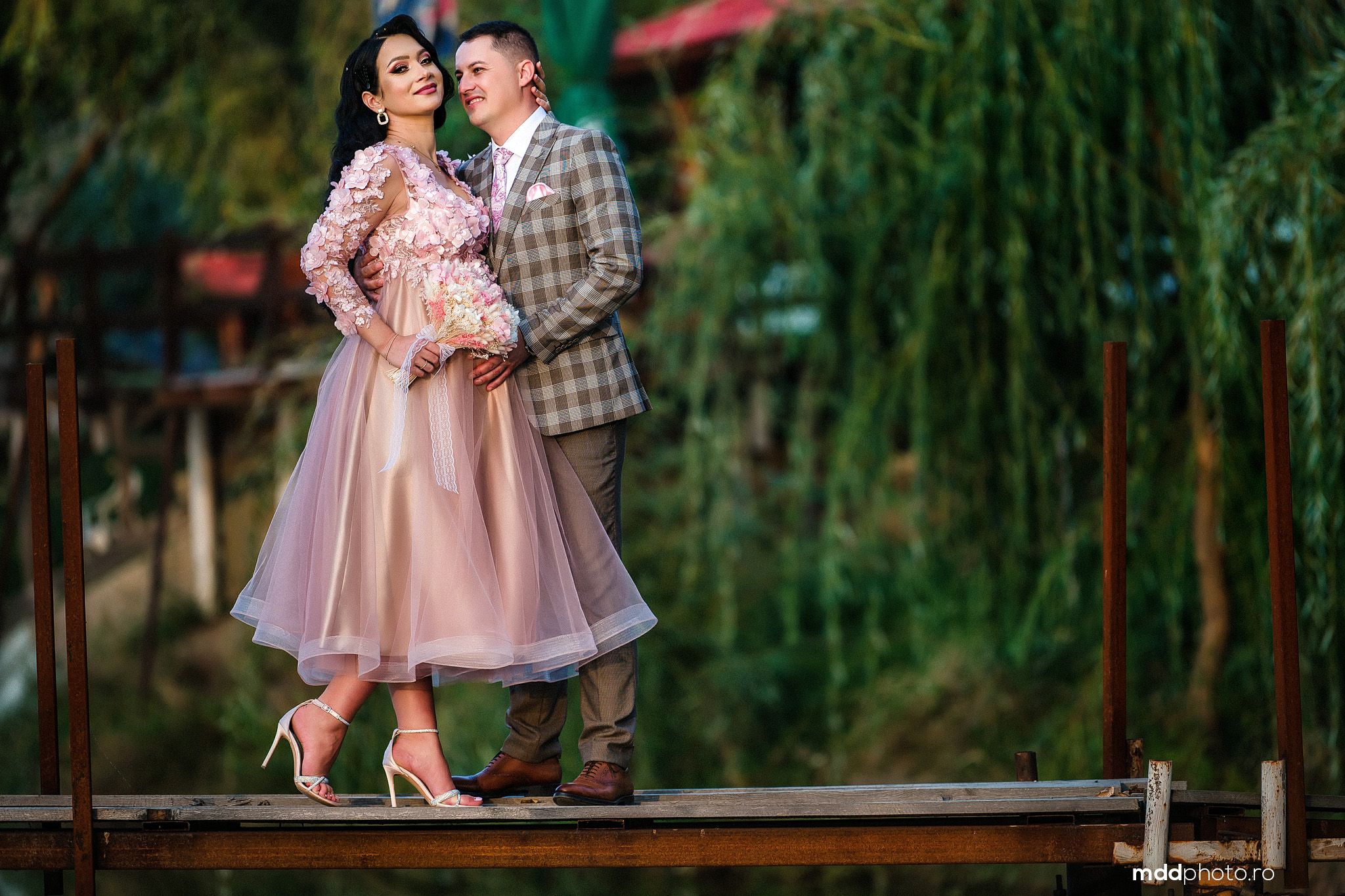 A couple embraces on a wooden deck with lush willow trees in the background.