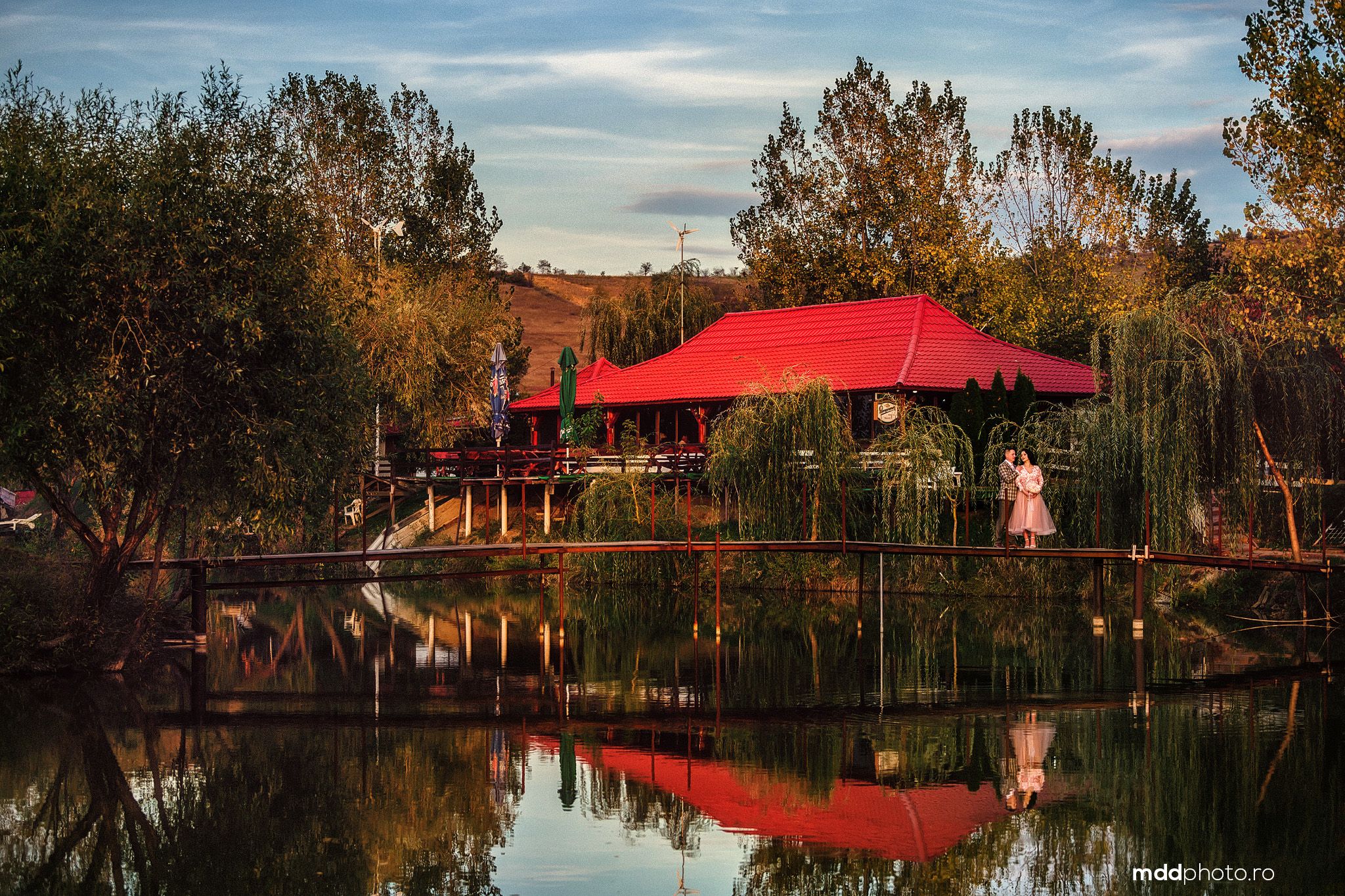 A rustic cabin with a bright red roof reflects perfectly in a calm pond during autumn sunset.