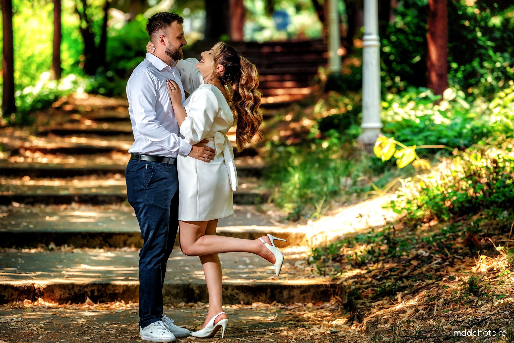 A romantic couple embraces on a sunlit forest path while standing among green foliage on a warm afternoon.