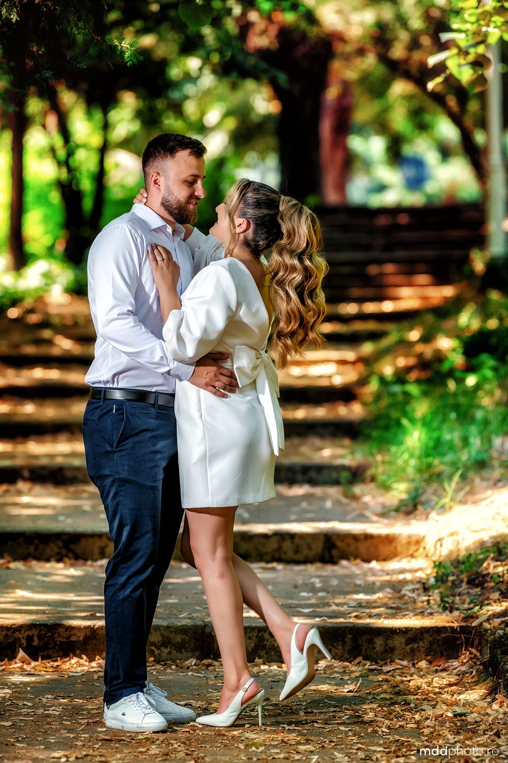 A romantic couple embraces on outdoor steps surrounded by lush greenery and trees in a park setting.