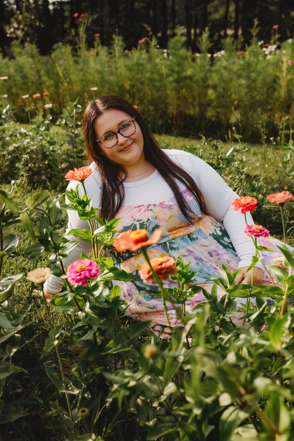 Young woman with glasses sitting in a lush garden surrounded by vibrant pink and orange zinnia flowers.