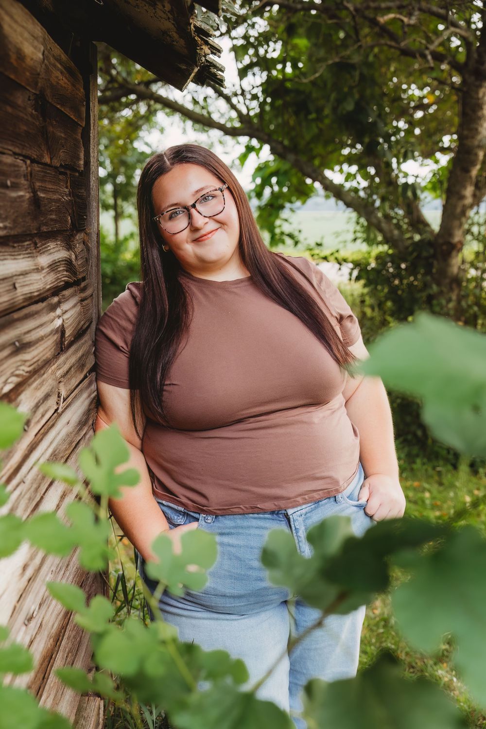 Young woman with glasses in brown tee and jeans leaning against rustic wooden wall surrounded by green foliage.