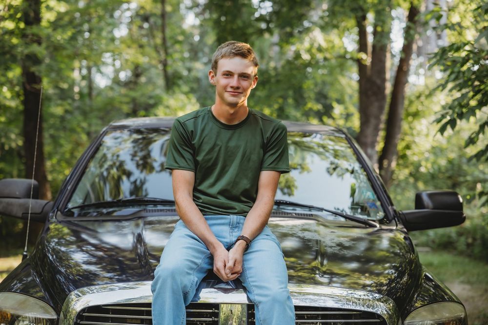 Young man in green t-shirt and jeans sitting on hood of black truck outdoors surrounded by lush green trees.