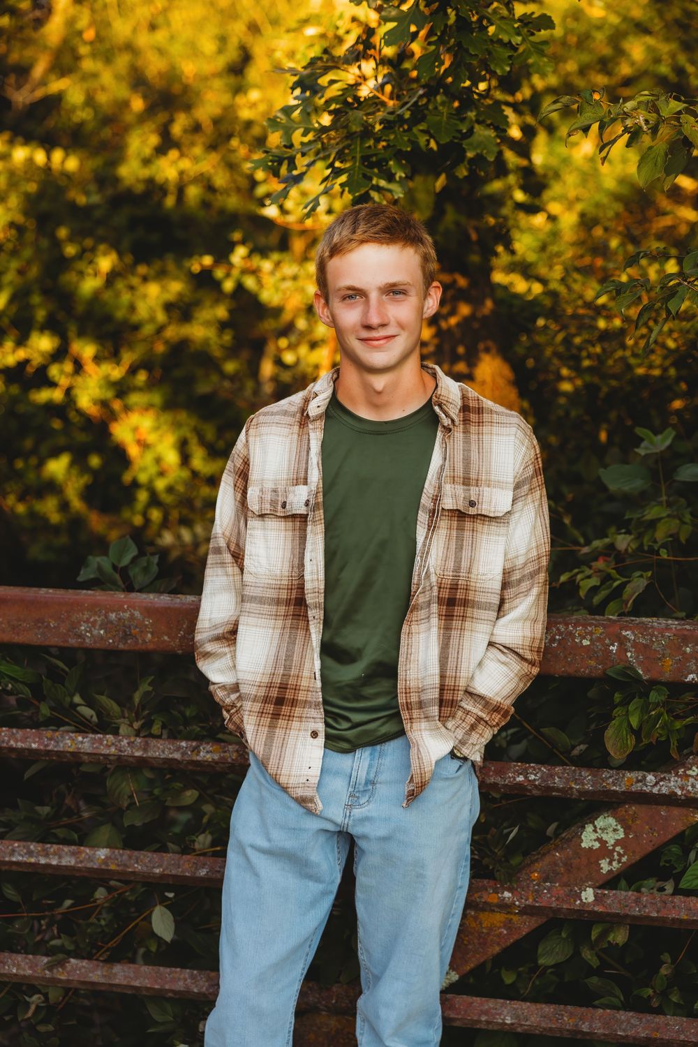 Young man in plaid flannel shirt and jeans smiling outdoors with autumn foliage background.