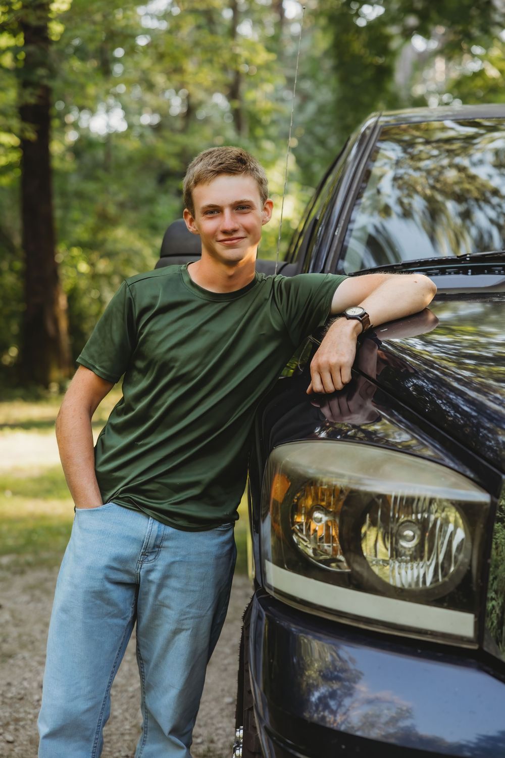 Young man in green t-shirt and jeans leaning on a dark truck in a wooded area, smiling confidently.