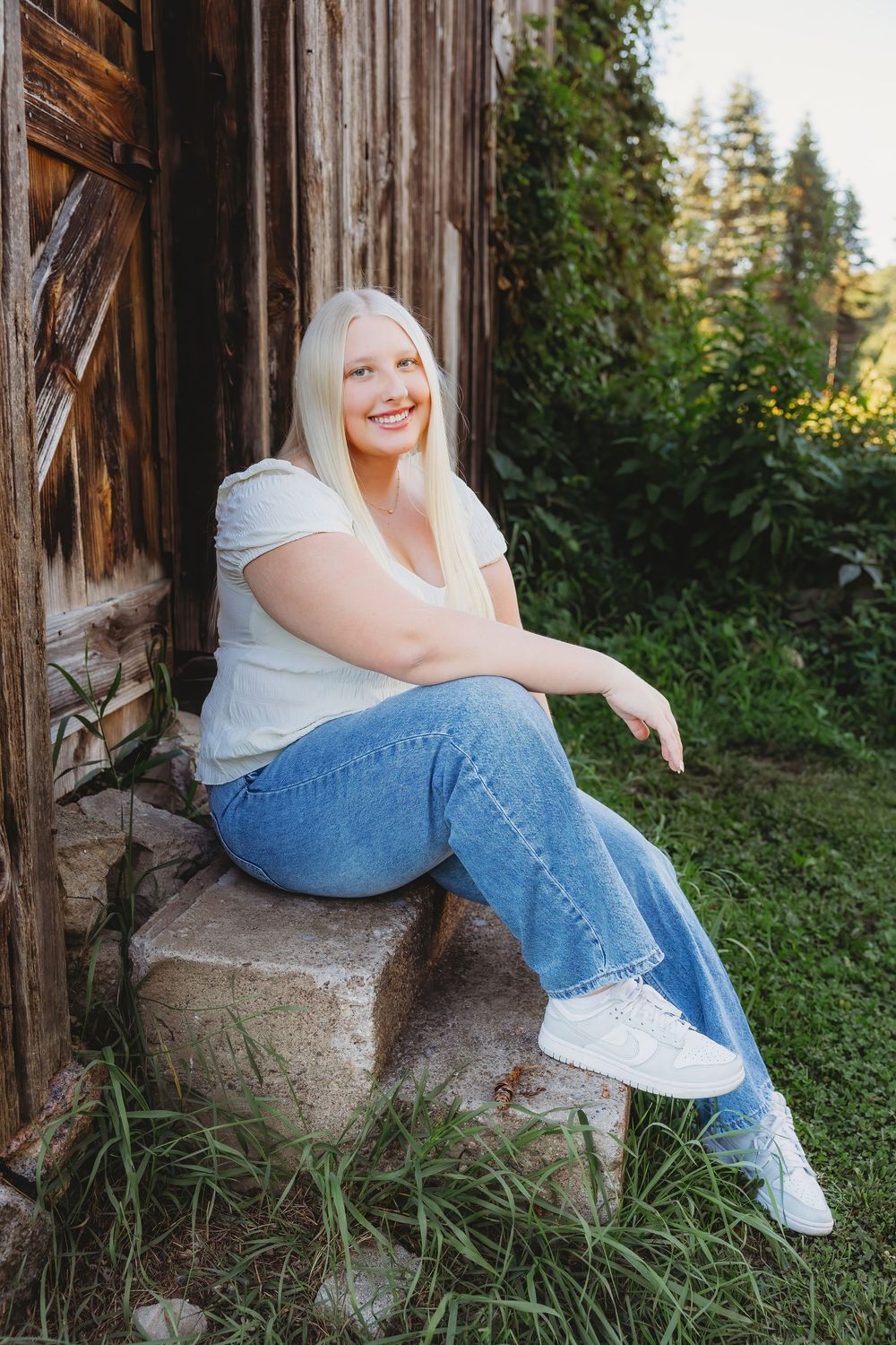 Smiling blonde woman sitting on stone steps beside a rustic wooden barn, wearing white tee and blue jeans outdoors.