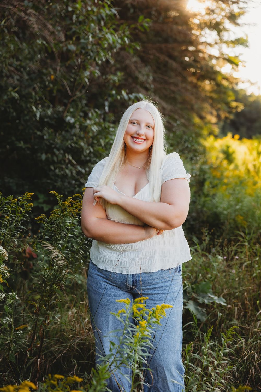 Smiling blonde woman in white top and jeans standing outdoors among wildflowers in golden hour sunlight.