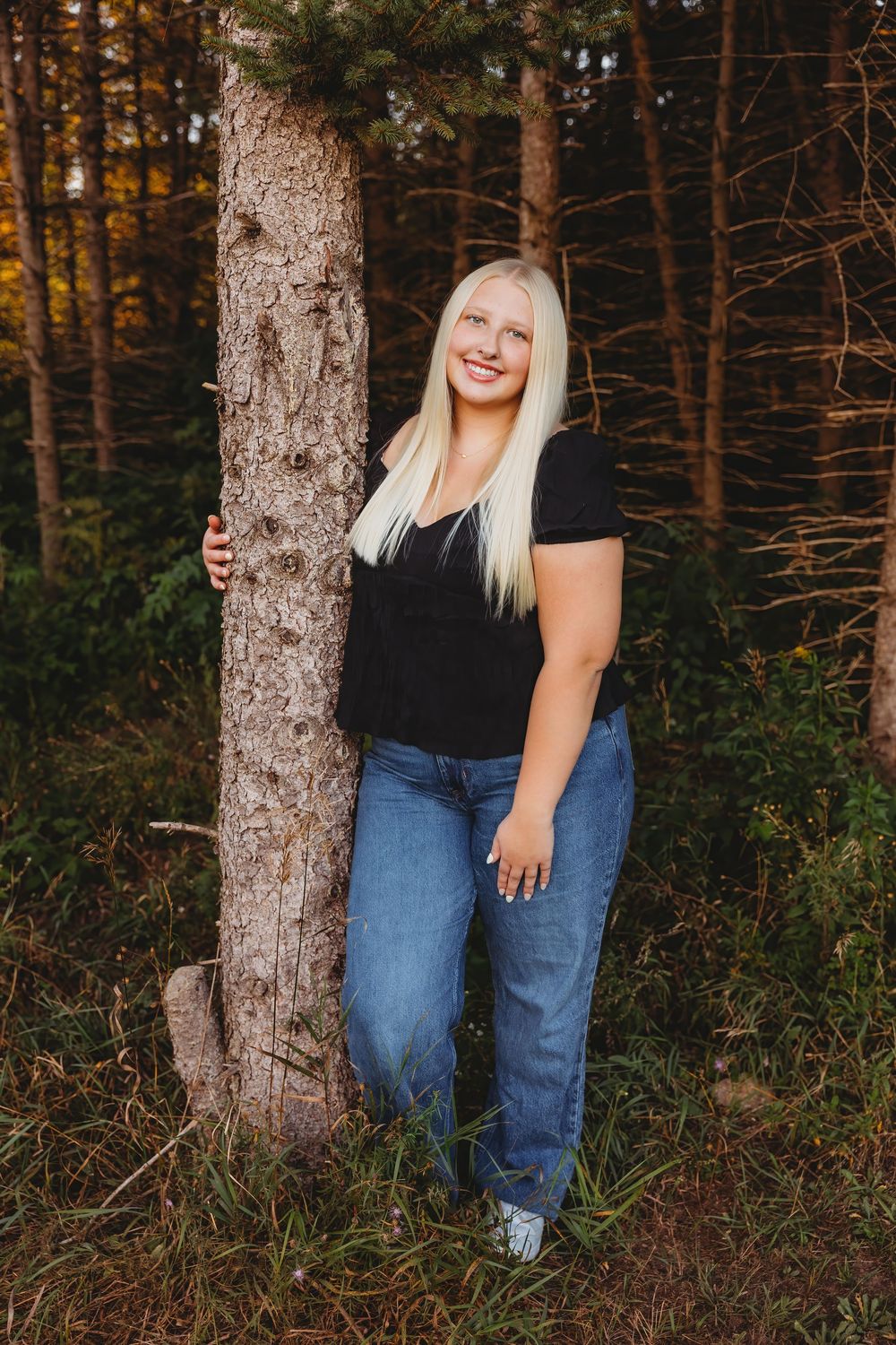 Smiling blonde woman in black top and jeans leaning against a tree in a forest setting.