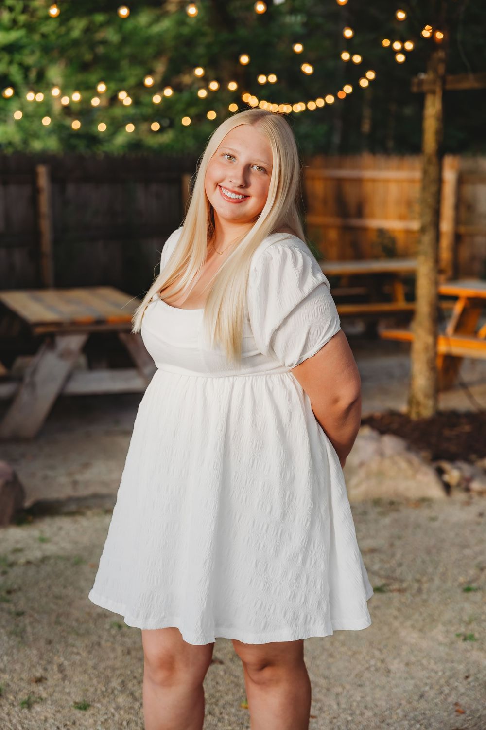 Young blonde woman in white dress smiling outdoors with string lights and wooden fence backdrop.