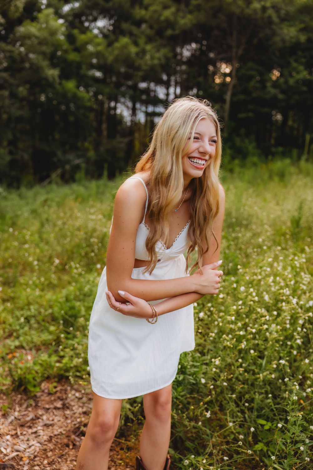 Young blonde woman in white sundress laughing joyfully outdoors in a wildflower meadow with forest background.