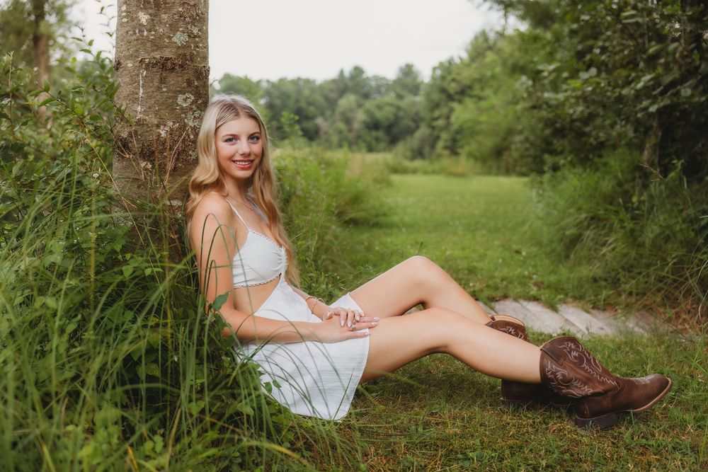 Young blonde woman in white dress and cowboy boots sitting against a tree in a lush green outdoor setting.