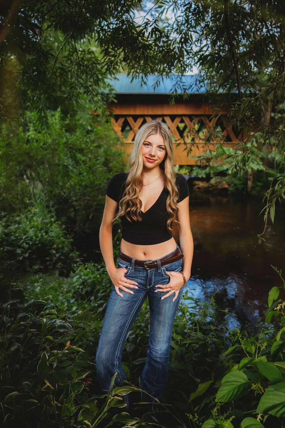 Smiling blonde woman in black crop top and jeans poses near a covered bridge surrounded by lush green trees.