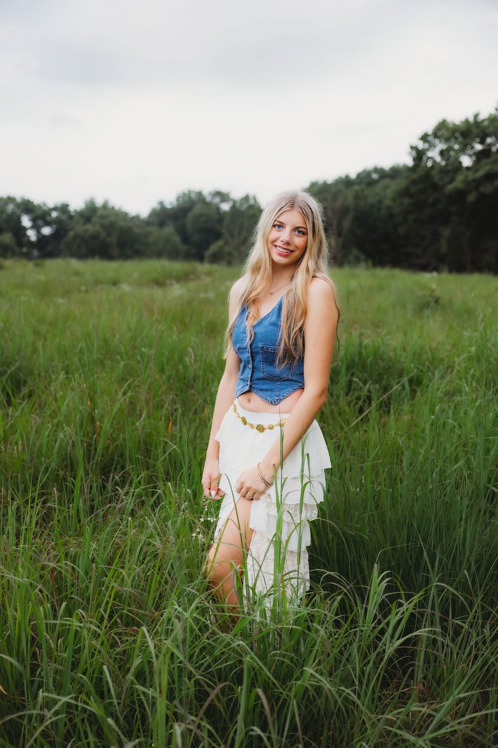 Smiling blonde woman in denim top and white skirt standing in tall green grass field.