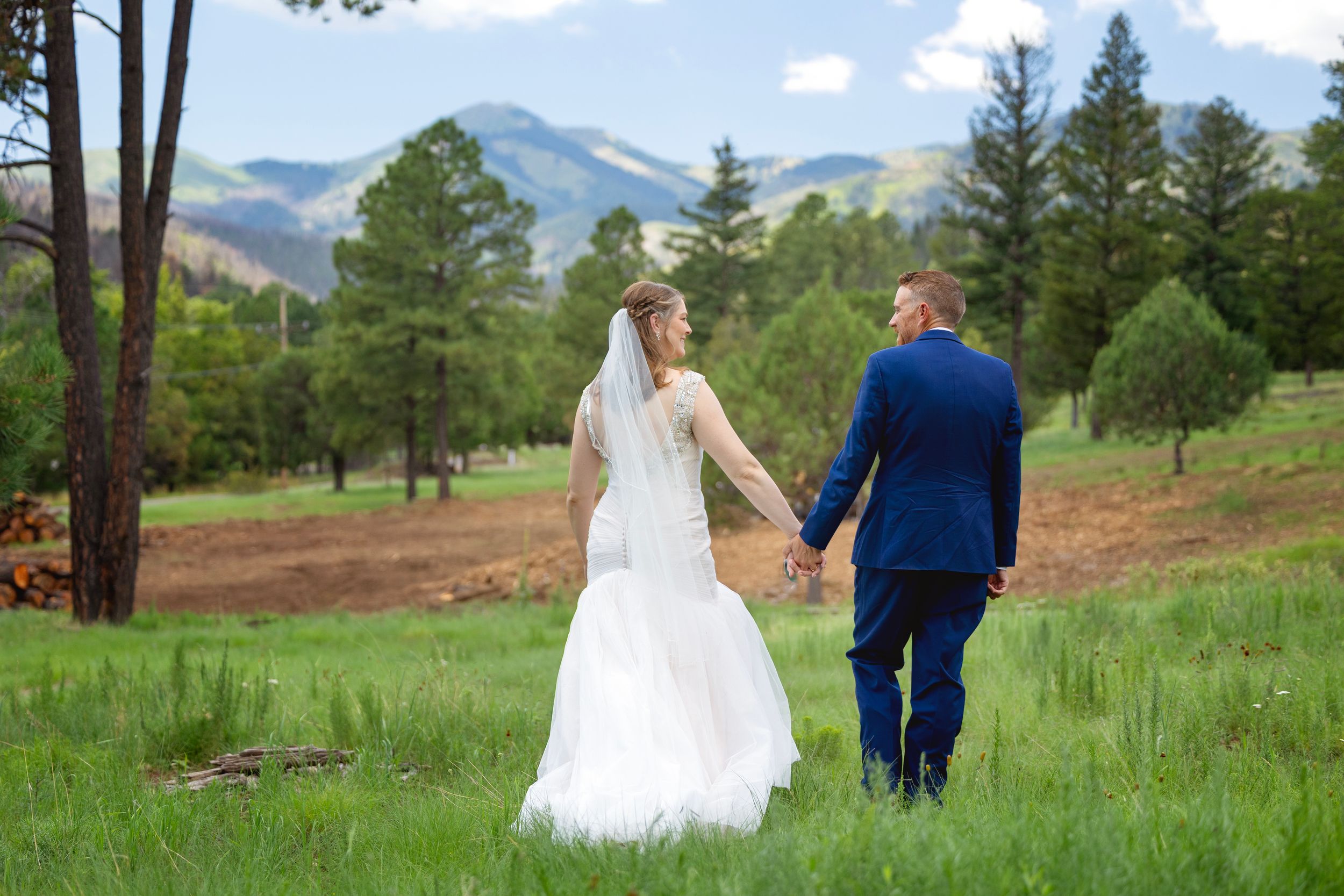 Bride and groom walking hand in hand through a green meadow with mountain views behind them in Ruidoso, NM.