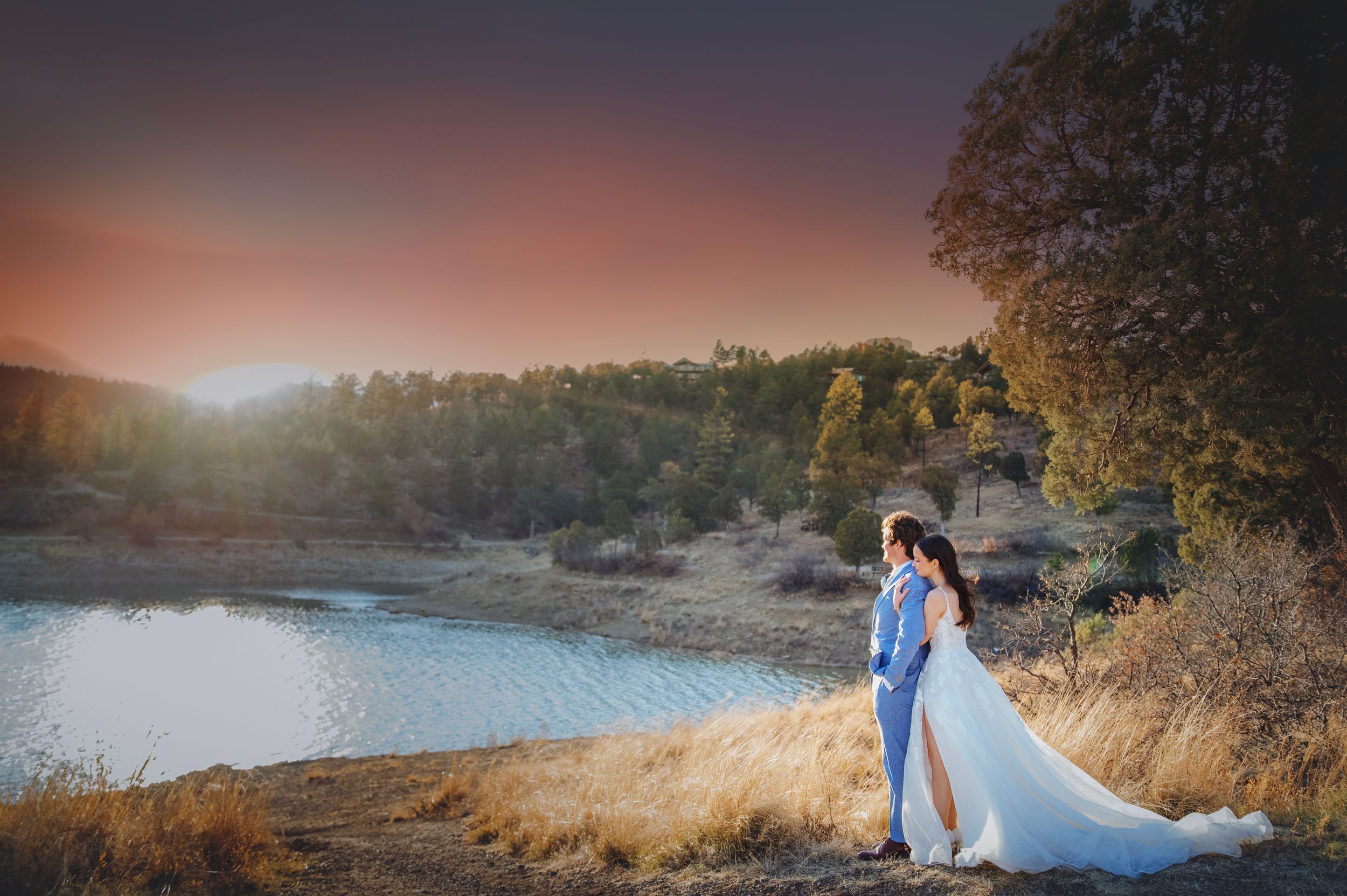 Couple embracing near a lake at sunset with warm golden light and rolling hills behind them.