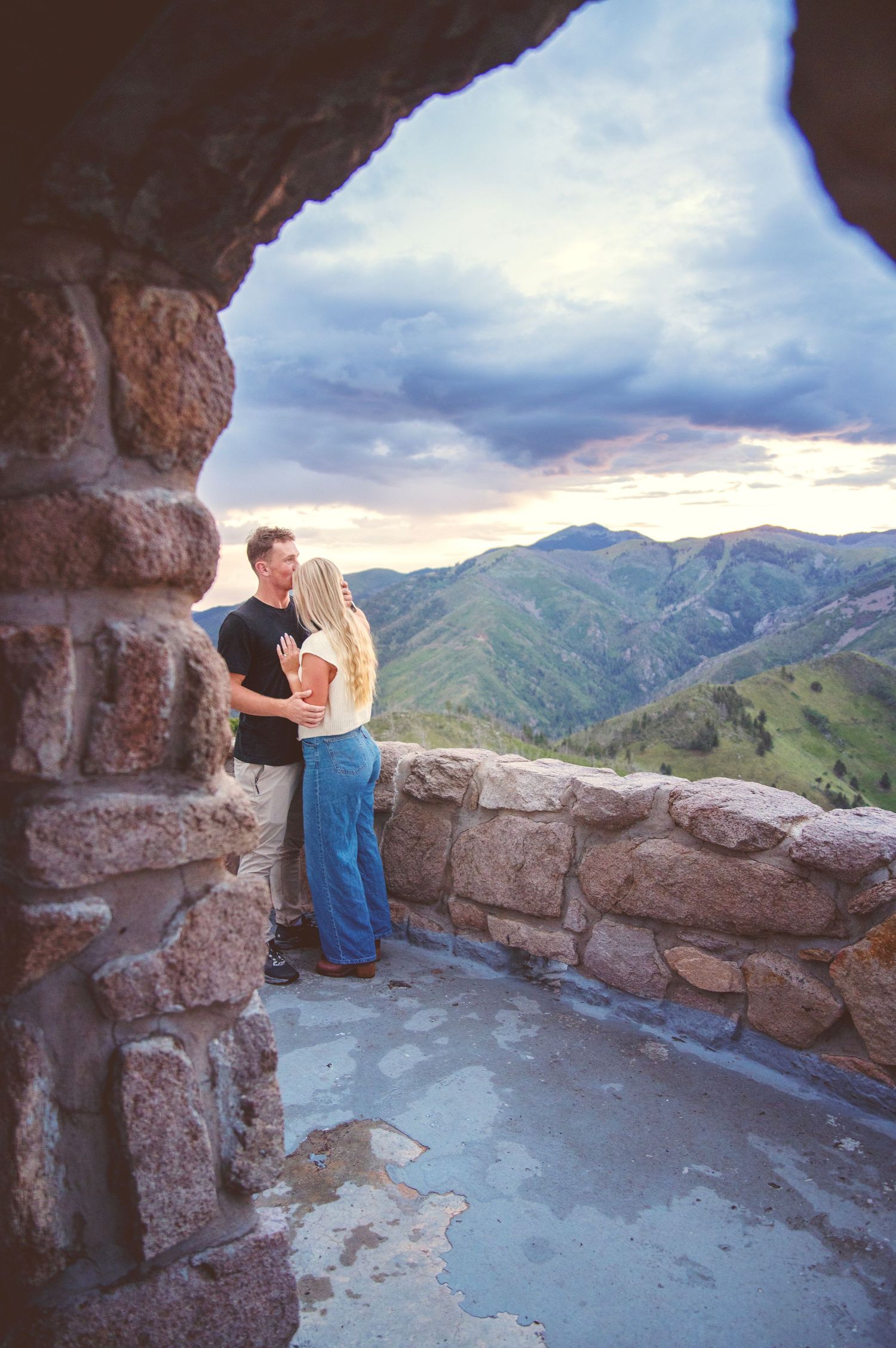 Couple kissing on a stone overlook with dramatic mountain and cloudy sky views behind them in Ruidoso, NM.