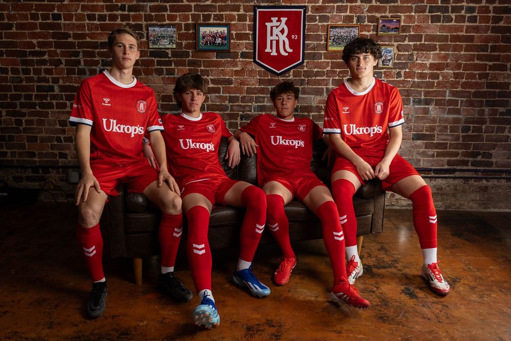 Soccer team members in red jerseys pose casually on wooden benches against a brick backdrop.
