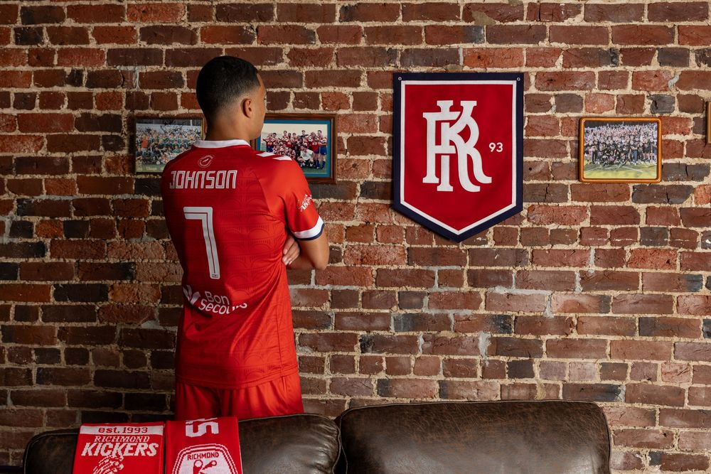 Soccer players in red uniforms sit against a rustic brick wall with team logos and banners.