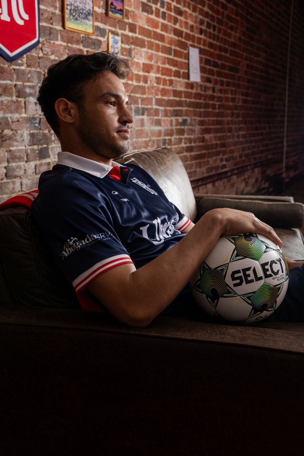 Soccer players holding Select brand balls while resting against brick wall in navy uniforms.