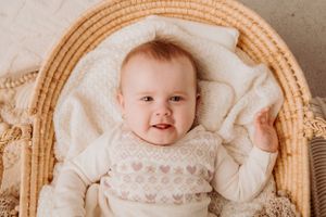 Baby in white lace dress smiling while sitting in wicker chair against neutral background.