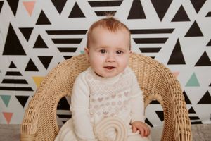Baby in cream-colored outfit sits in vintage wicker chair against geometric pattern backdrop.