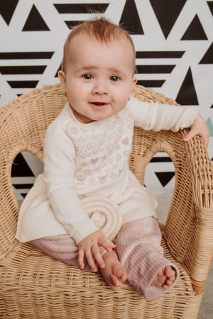 Smiling baby in delicate white outfit poses in wicker chair against modern geometric wall.