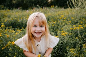 Child with blonde hair smiling brightly in a summer meadow filled with yellow flowers.