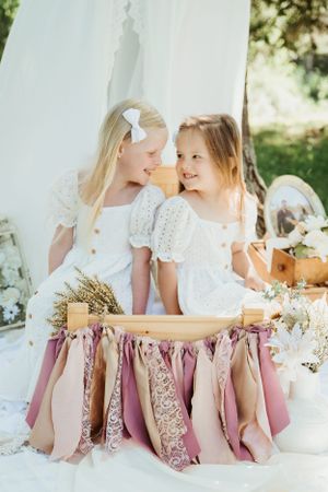 Two children in white dresses share sweet moment at vintage-styled outdoor tea party.